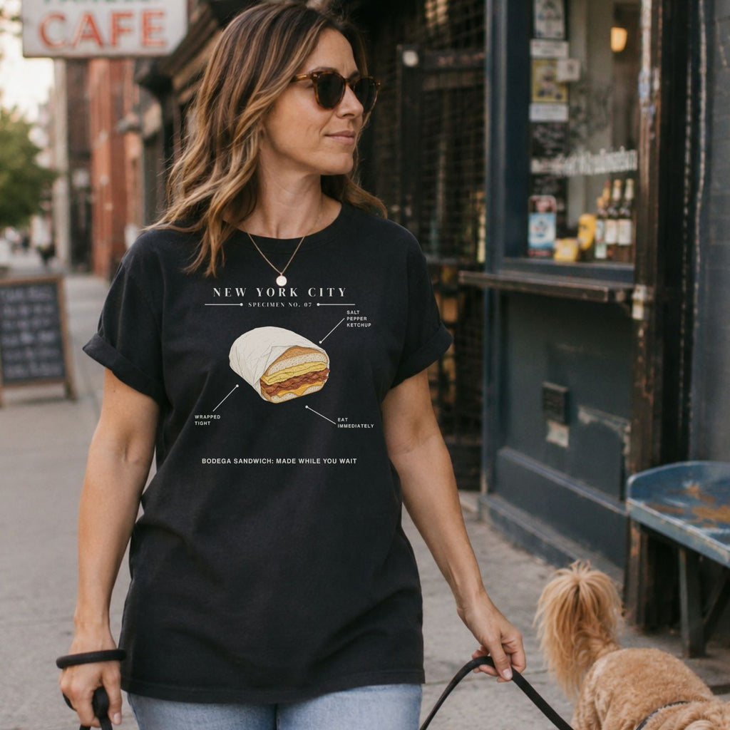 Woman walking a dog on a sidewalk wearing a Black t-shirt with a graphic of a bodega sandwich and text. 