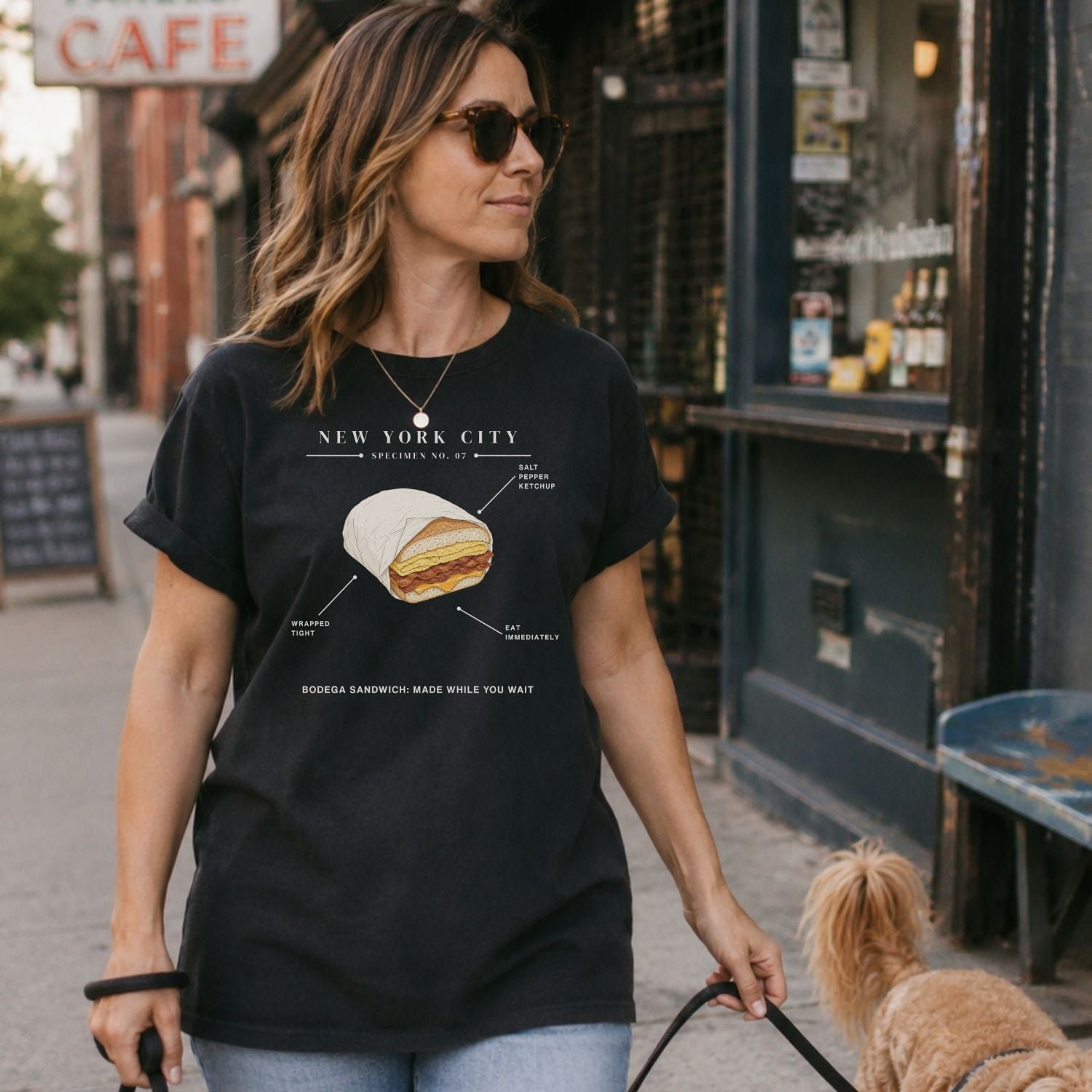 Woman walking a dog on a sidewalk wearing a Black t-shirt with a graphic of a bodega sandwich and text. 