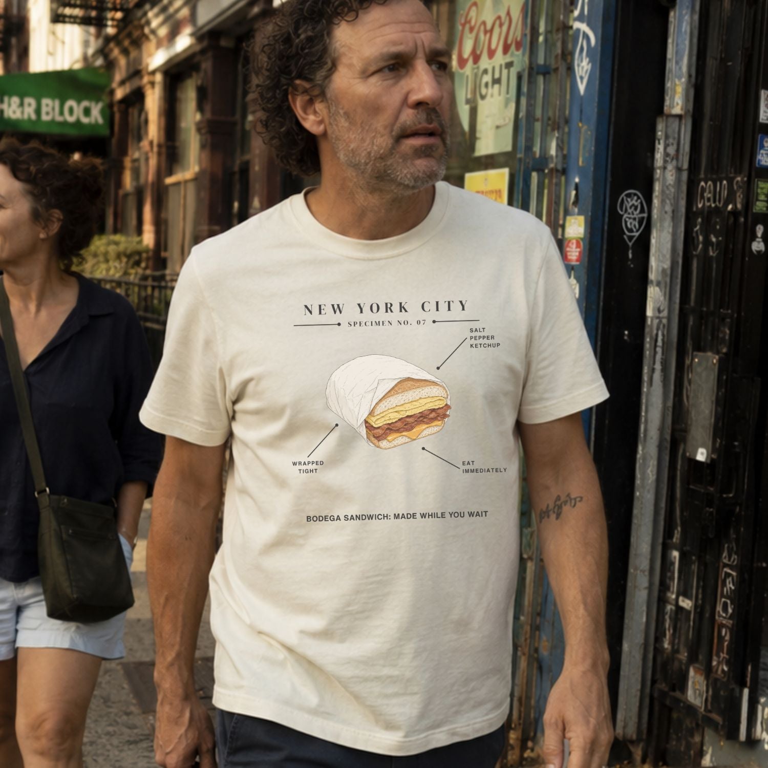 A man wearing an Ivory Comfort Colors t-shirt featuring a New York City bodega sandwich graphic with specimen-style callouts and “Bodega Sandwich: Made While You Wait” text.