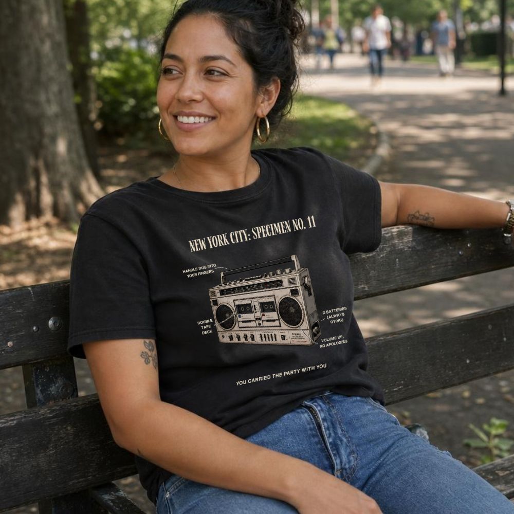 Woman wearing a black Comfort Colors t-shirt with a vintage boombox graphic design, sitting on a bench in an outdoor setting.