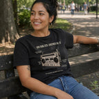Woman wearing a black Comfort Colors t-shirt with a vintage boombox graphic design, sitting on a bench in an outdoor setting.