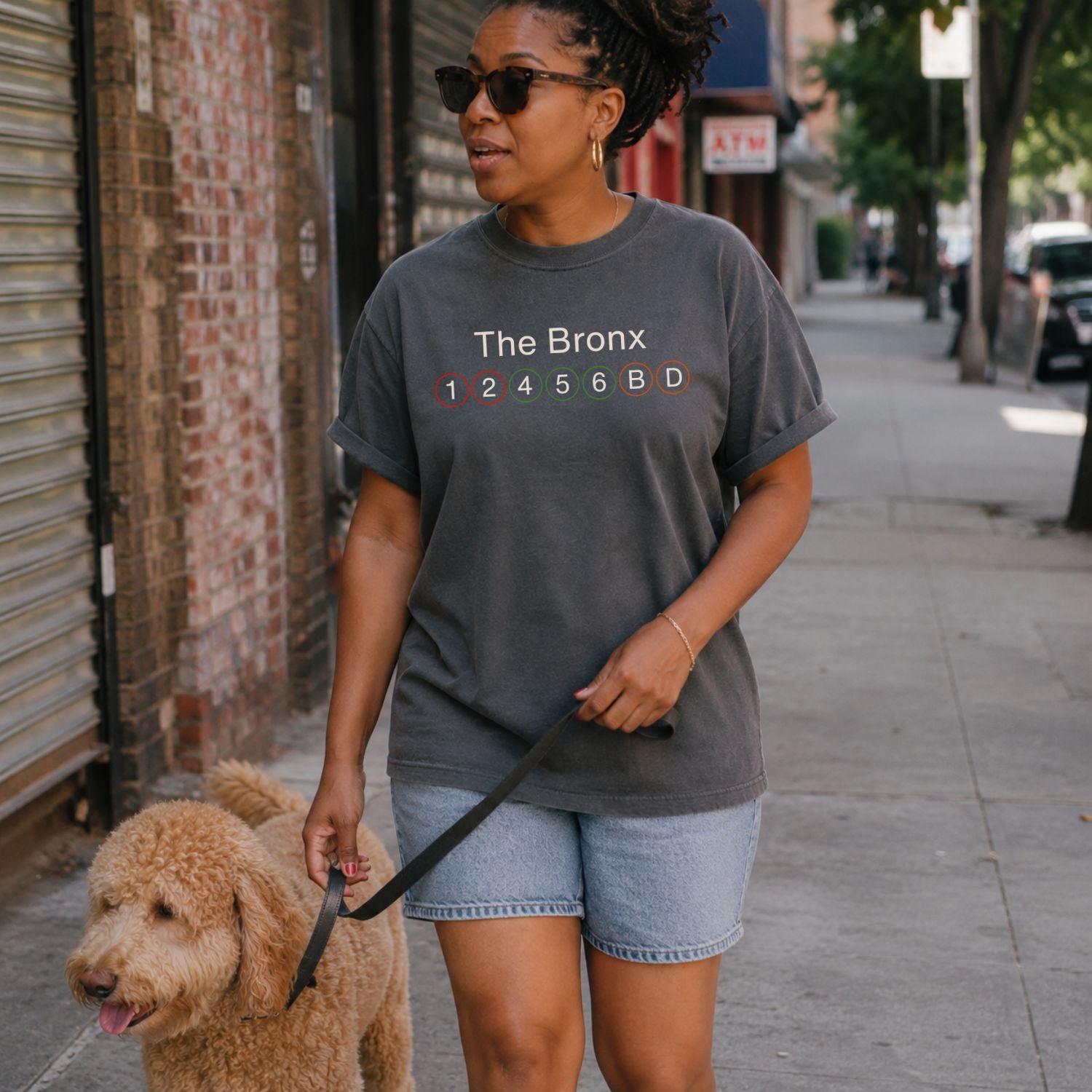 Woman walking a dog on a city street wearing a Pepper colored t-shirt with 'The Bronx' and subway line numbers. 