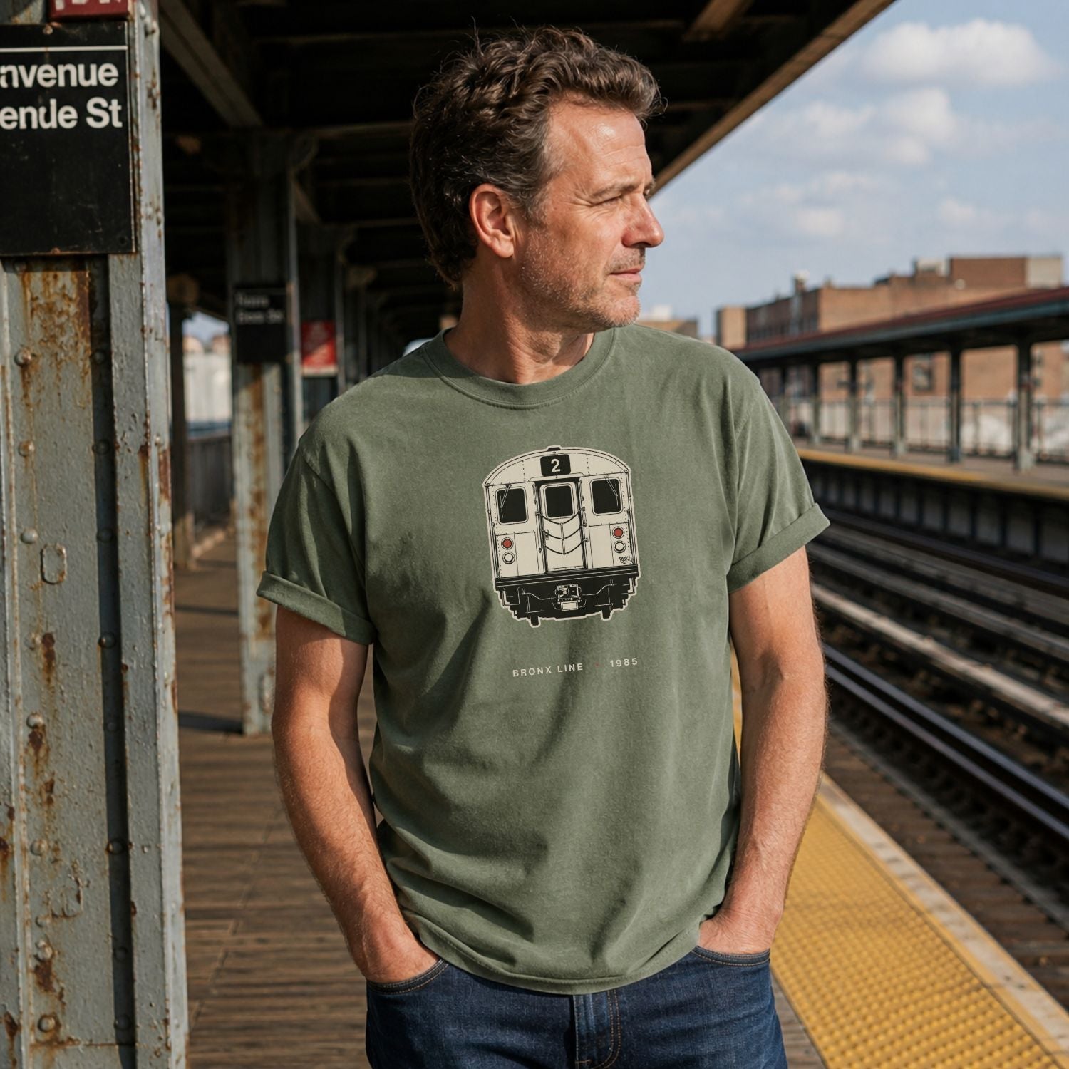 A man wearing a Moss Green t-shirt with a NYC subway train graphic while standing on an elevated station platform. 