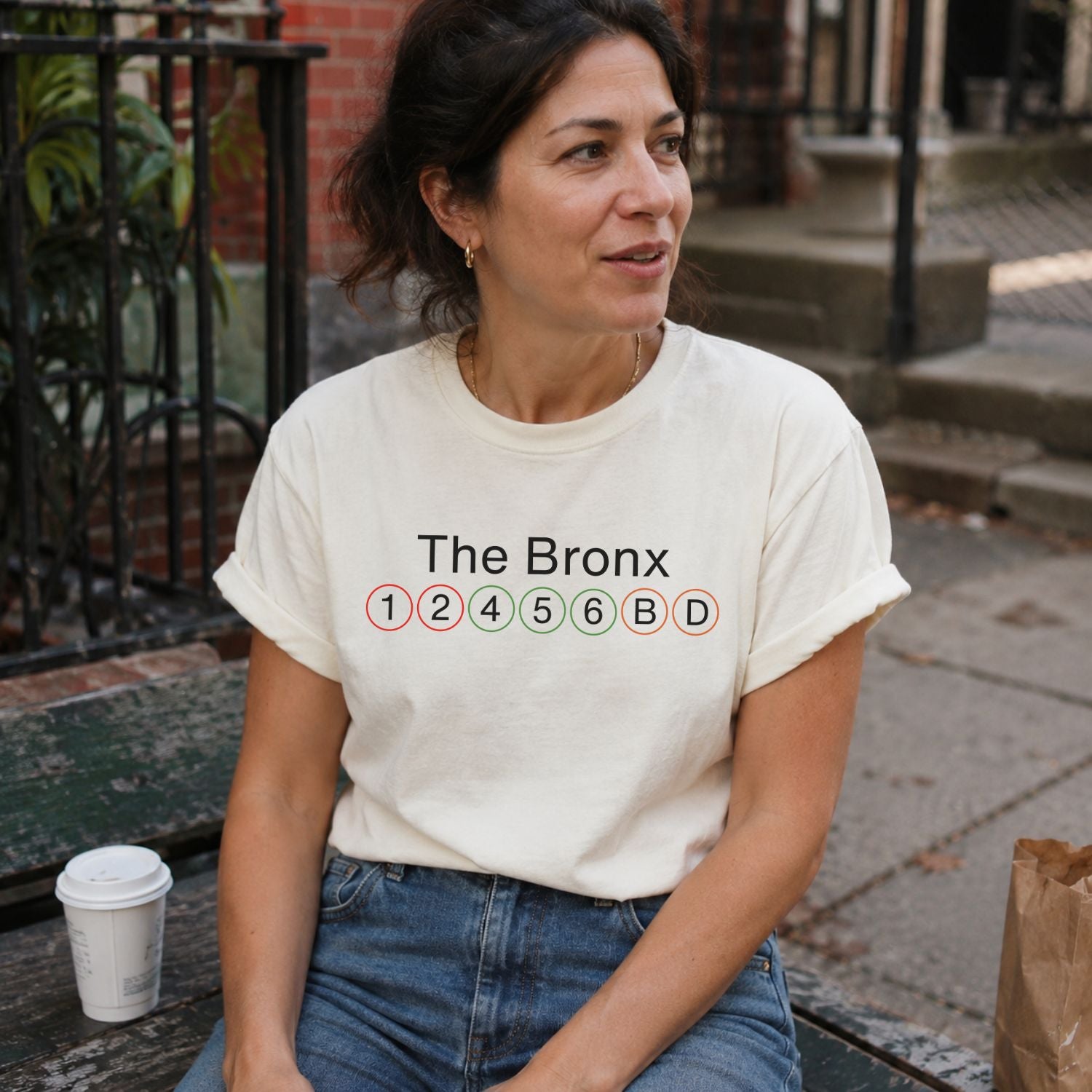 Woman wearing an Ivory Comfort Colors t-shirt featuring a minimalist Bronx subway line graphic with The Bronx text and classic NYC train icons sitting outdoors.