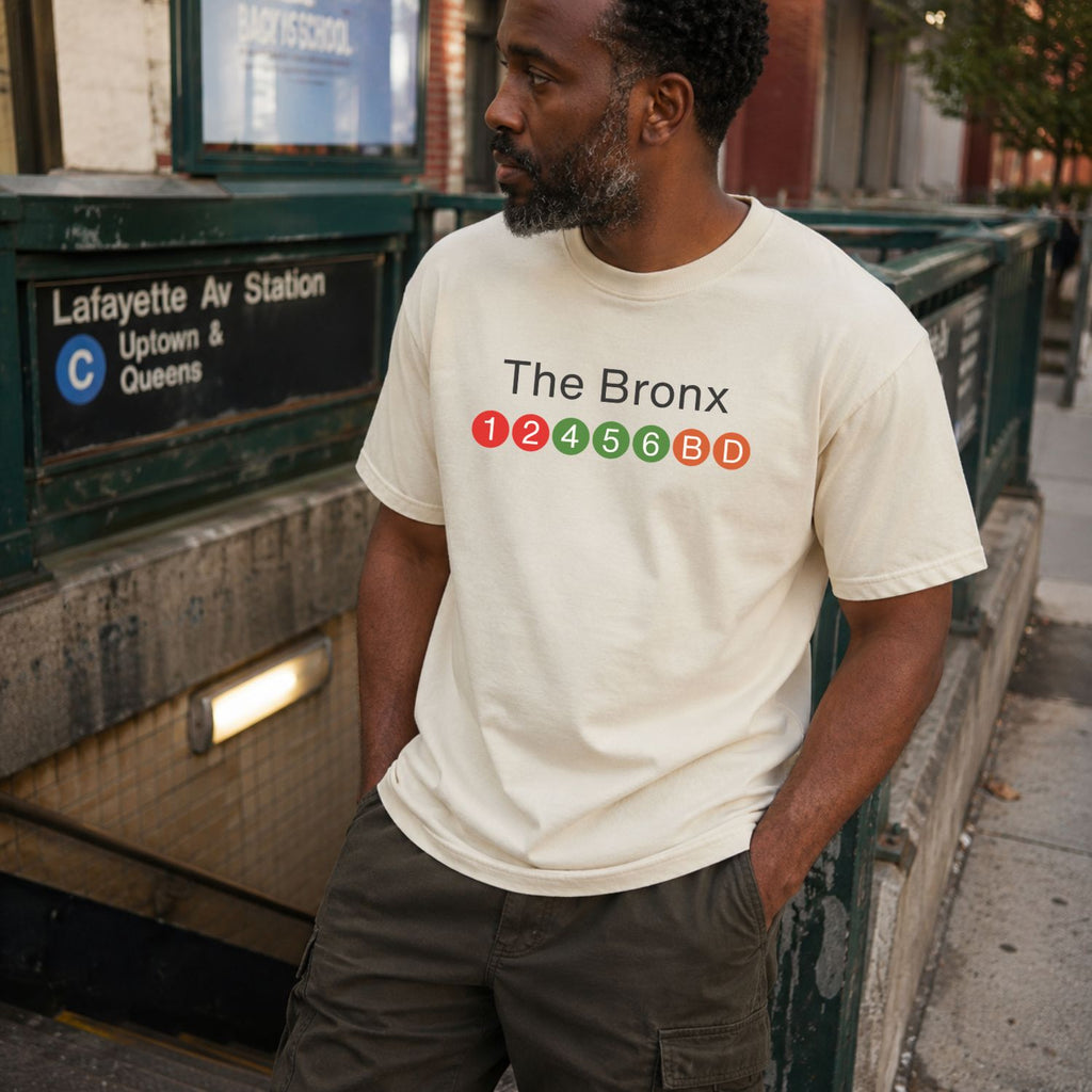 Man wearing an Ivory t-shirt with 'The Bronx' text and NYC subway lines  standing near a subway station sign.