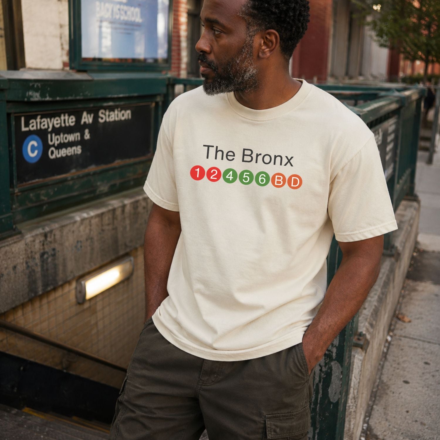 Man wearing an Ivory t-shirt with 'The Bronx' text and NYC subway lines  standing near a subway station sign.