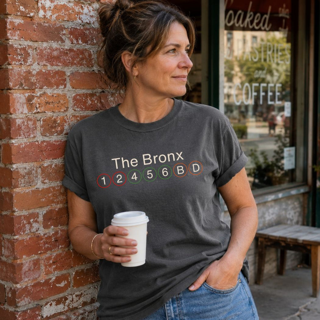 Woman wearing a Pepper colored Comfort Colors t-shirt with 'The Bronx' and subway line text, holding a coffee cup, standing against a brick wall.