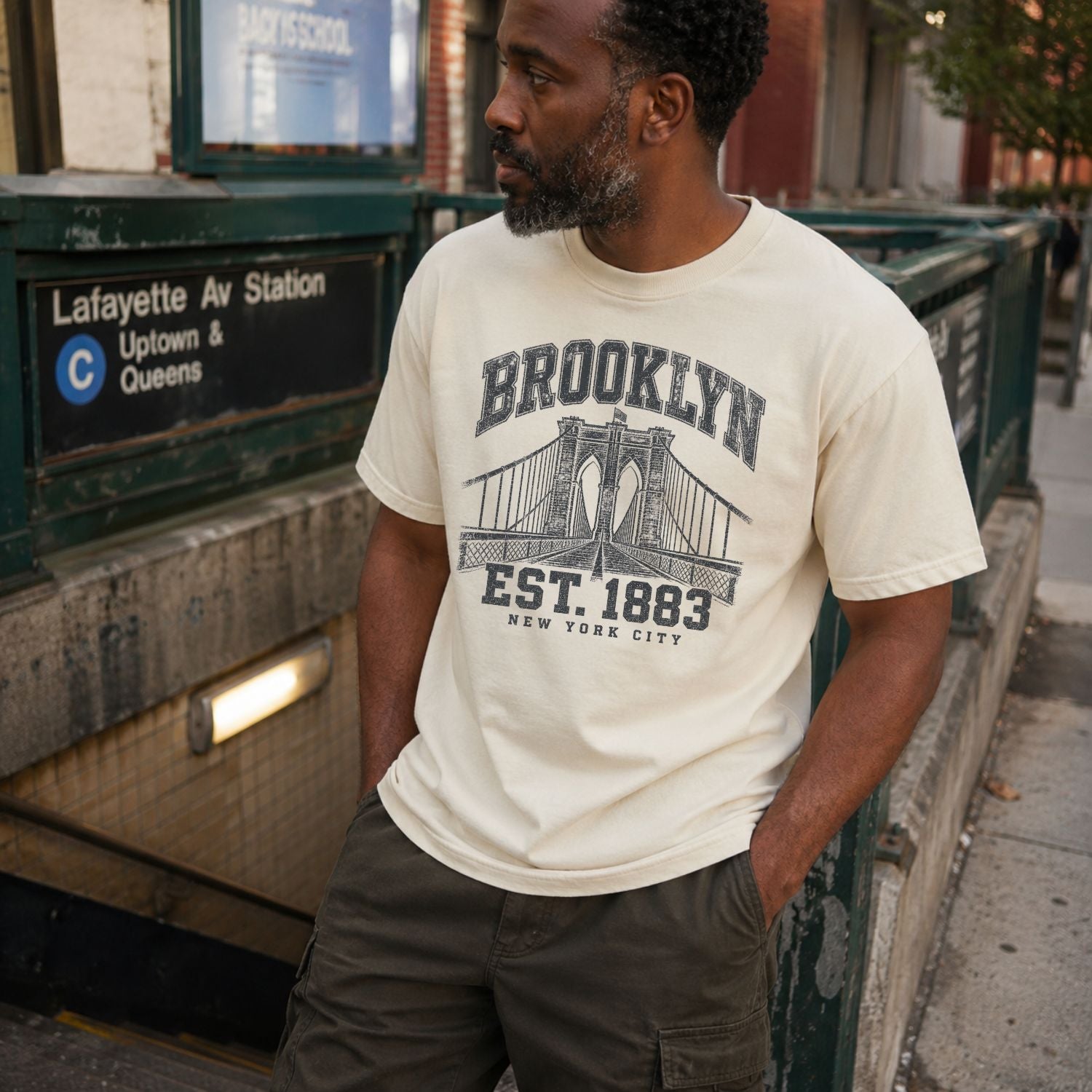 Man wearing an Ivory Vintage-style Comfort Colors t-shirt, featuring the Brooklyn Bridge and “Brooklyn Est. 1883” text.standing near a subway station sign.