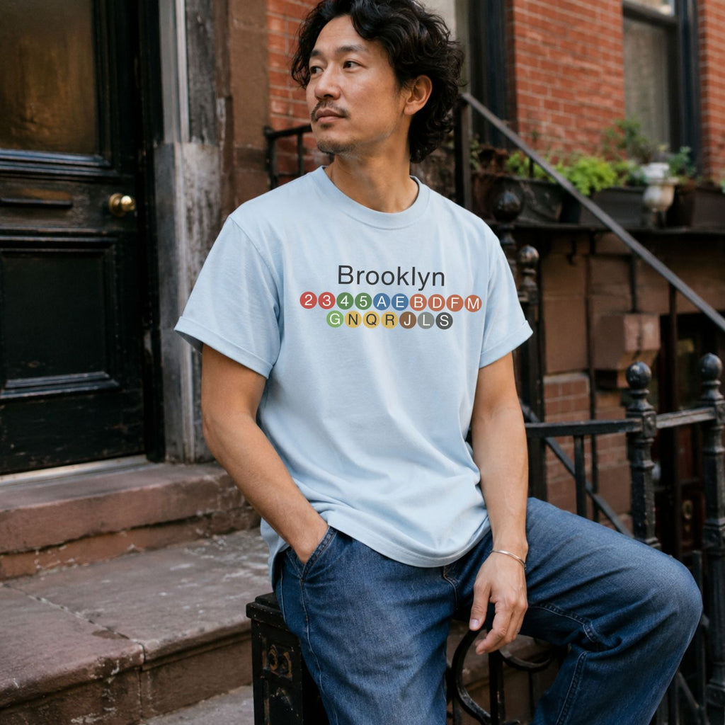 Man wearing a Light blue t-shirt with 'Brooklyn' and subway line numbers sitting on a stoop.