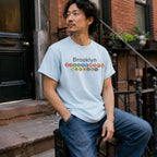 Man wearing a Light blue t-shirt with 'Brooklyn' and subway line numbers sitting on a stoop.