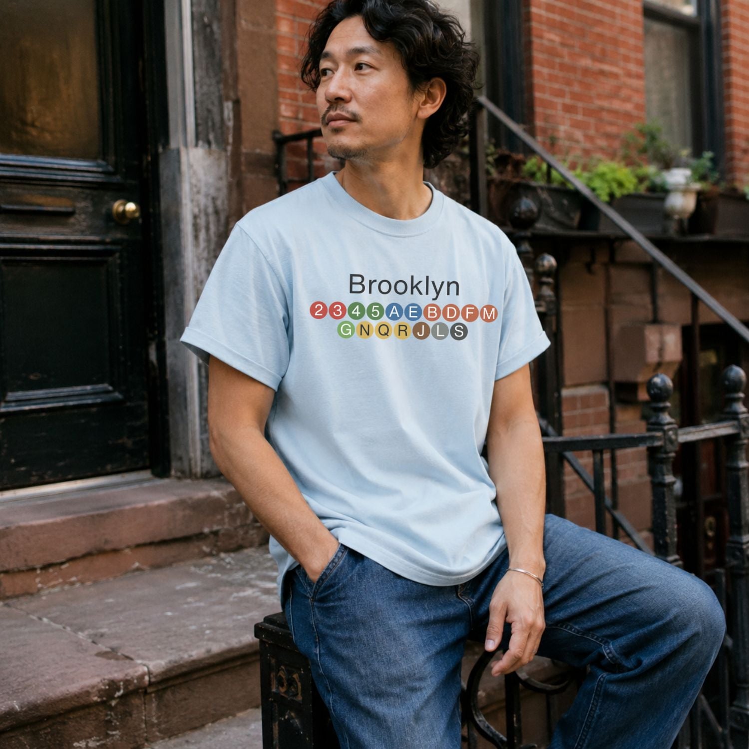 Man wearing a Light blue t-shirt with 'Brooklyn' and subway line numbers sitting on a stoop.