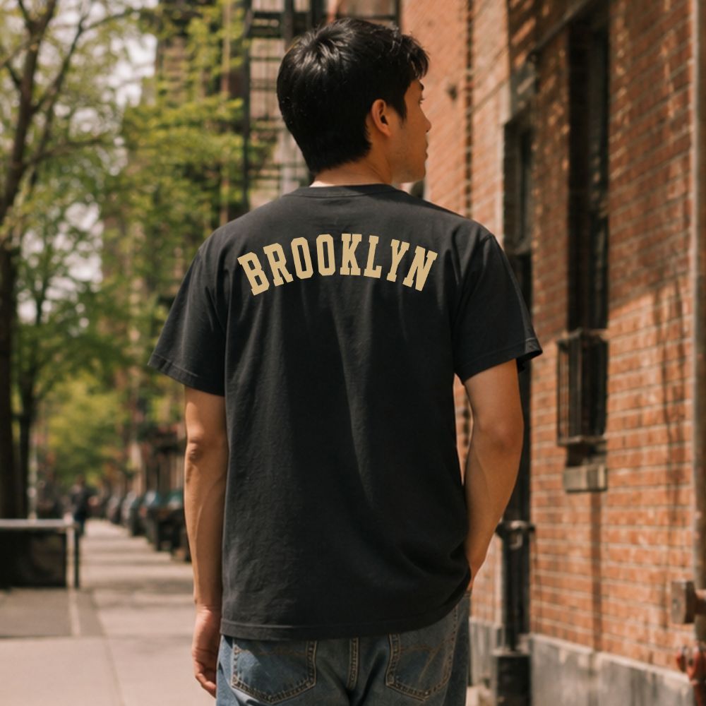 Person wearing a black Comfort Colors t-shirt with 'Brooklyn' printed on the back, standing on a street.