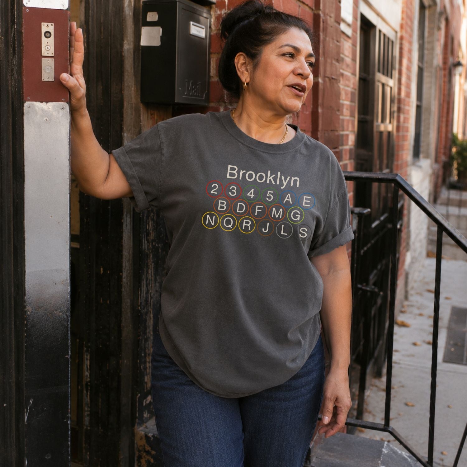 Woman wearing a Pepper colored Comfort Colors t-shirt featuring the word “Brooklyn” above colorful NYC subway line circles representing the subway routes that run through Brooklyn.