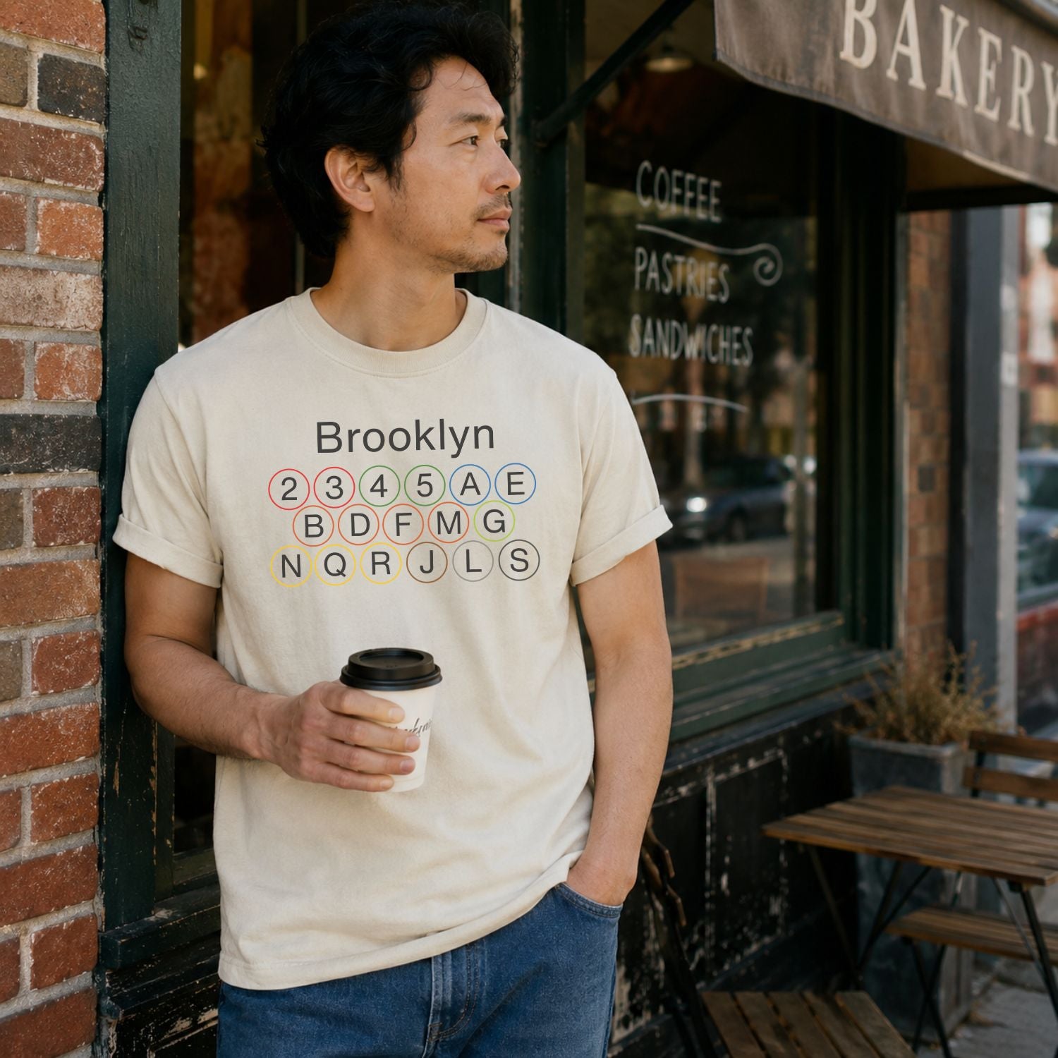 Man wearing an Ivory t-shirt with 'Brooklyn' and subway line numbers printed on it while holding a coffee cup outside a bakery.