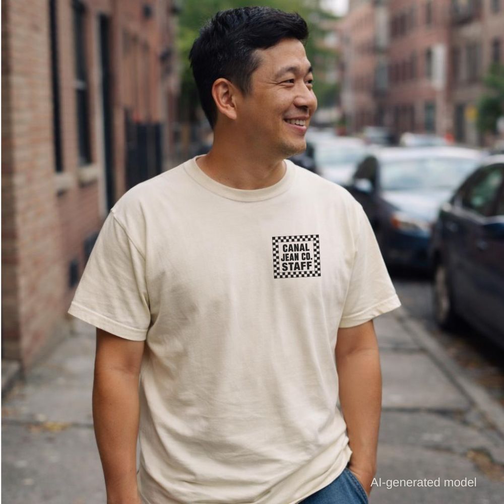 Man wearing an Ivory Comfort Colors T-shirt with black Canal Jean Co. NYC small front chest staff print, while standing on a NY sidewalk. 