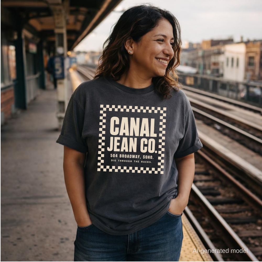 Woman wearing a pepper colored Comfort Colors T-shirt with ivory Canal Jean Co. NYC front print, while standing on a subway platform. 