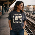 Woman wearing a pepper colored Comfort Colors T-shirt with ivory Canal Jean Co. NYC front print, while standing on a subway platform. 