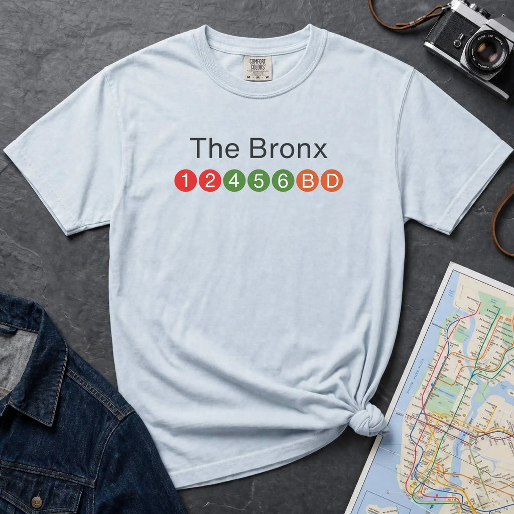 Light blue  t-shirt with 'The Bronx' text and NYC subway lines on a dark surface with a denim jacket, camera and a map.