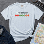 Light blue  t-shirt with 'The Bronx' text and NYC subway lines on a dark surface with a denim jacket, camera and a map.