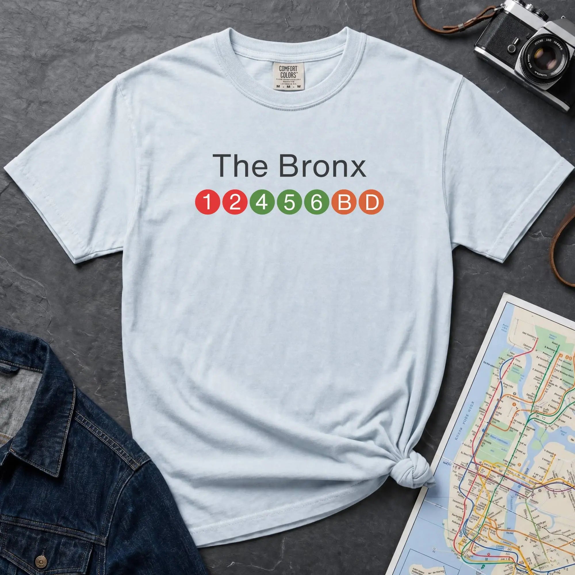Light blue  t-shirt with 'The Bronx' text and NYC subway lines on a dark surface with a denim jacket, camera and a map.