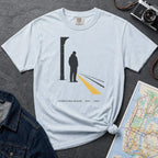 Light blue t-shirt with a graphic design of a person on a train platform, placed on a dark surface with a map and denim jacket.