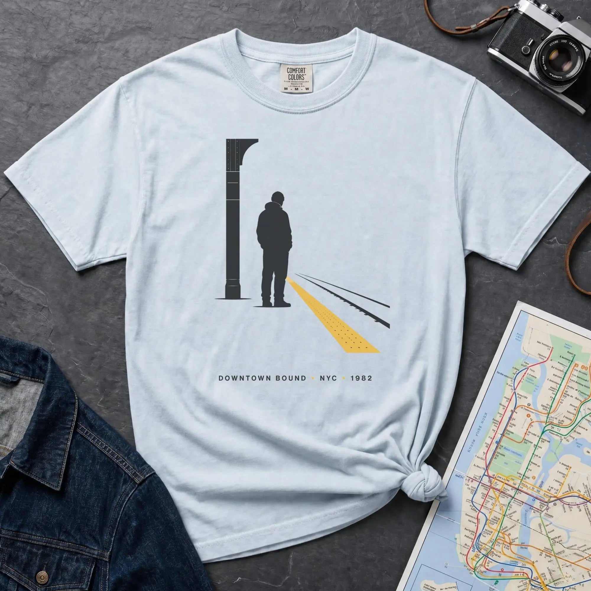 Light blue t-shirt with a graphic design of a person on a train platform, placed on a dark surface with a map and denim jacket.