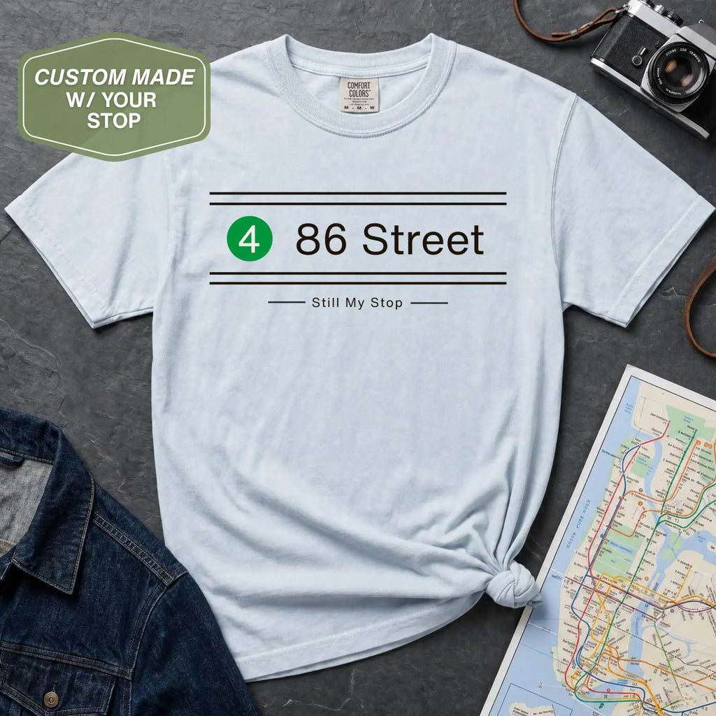 Light blue t-shirt with '86 Street' design on a gray surface with map and camera.