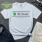 Light blue t-shirt with '86 Street' design on a gray surface with map and camera.