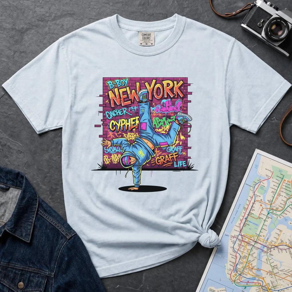 Light blue t-shirt with a colorful graphic design featuring a breakdancer and text on a dark surface with a denim jacket and a map.