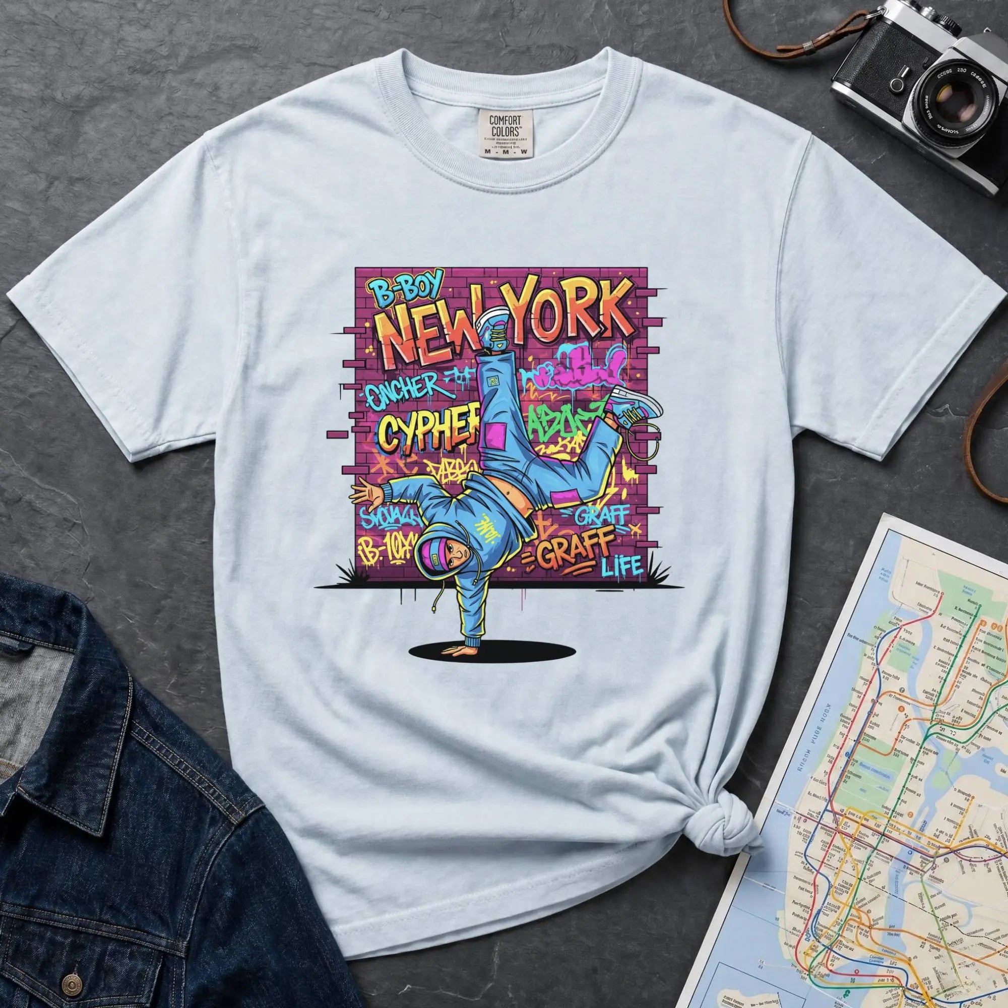 Light blue t-shirt with a colorful graphic design featuring a breakdancer and text on a dark surface with a denim jacket and a map.