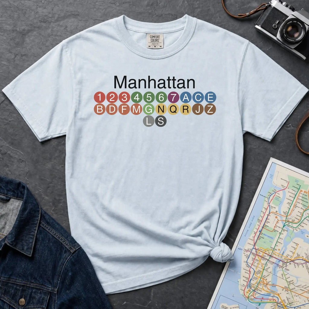 Light blue Comfort Colors t-shirt featuring a minimalist Manhattan subway line graphic with colorful NYC train icons.