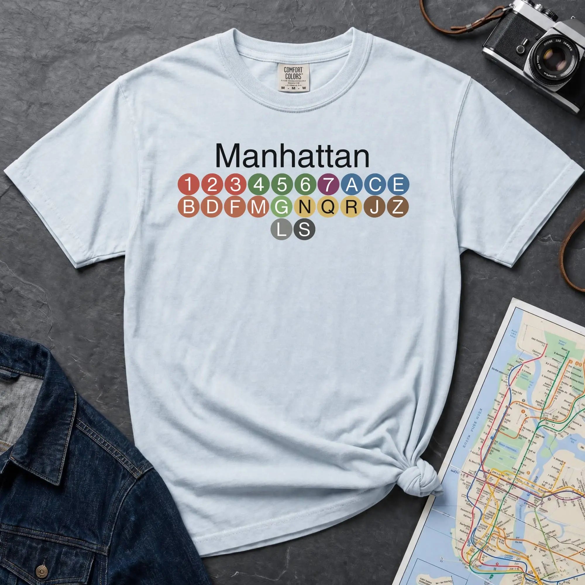 Light blue Comfort Colors t-shirt featuring a minimalist Manhattan subway line graphic with colorful NYC train icons.