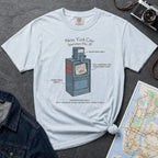 Light blue t-shirt with a New York City newspaper box design on a gray surface with a map and camera.