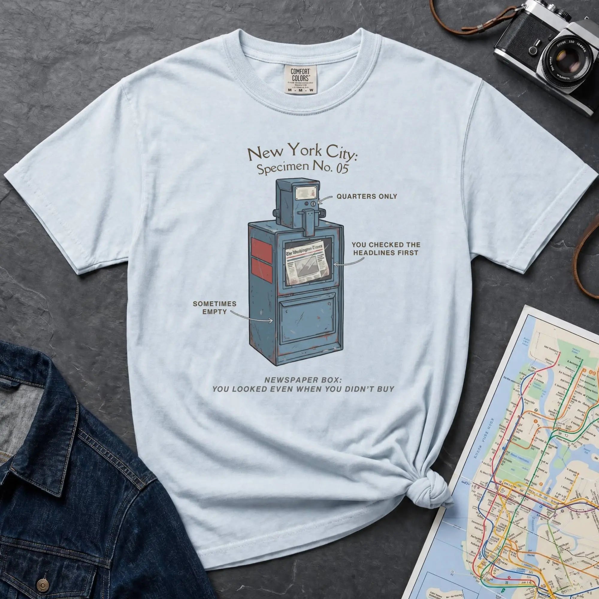 Light blue t-shirt with a New York City newspaper box design on a gray surface with a map and camera.