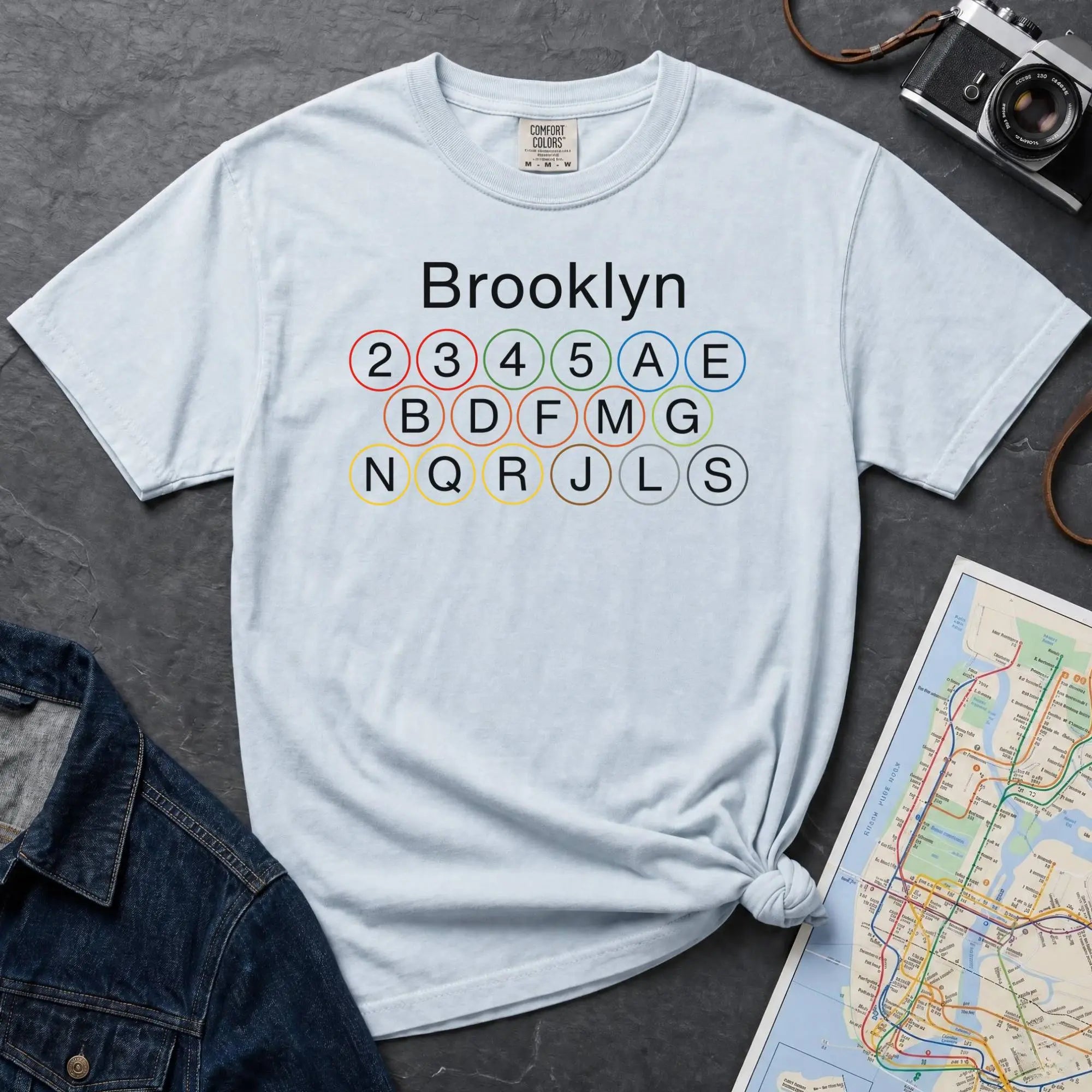 Light blue t-shirt with 'Brooklyn' and subway line numbers printed on it, placed on a dark surface with a map and denim jacket.
