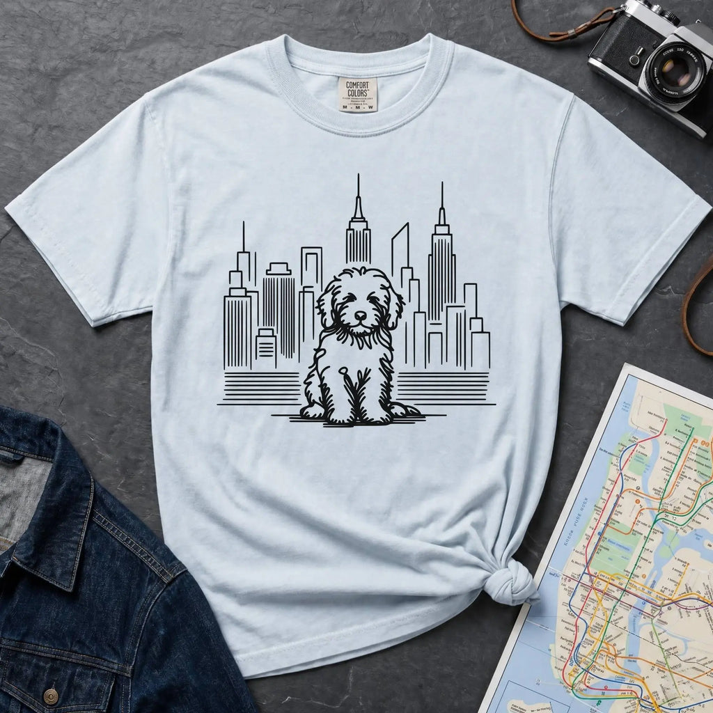 Light blue t-shirt with a Golden Doodle dog and cityscape design on a gray surface with a map and camera.