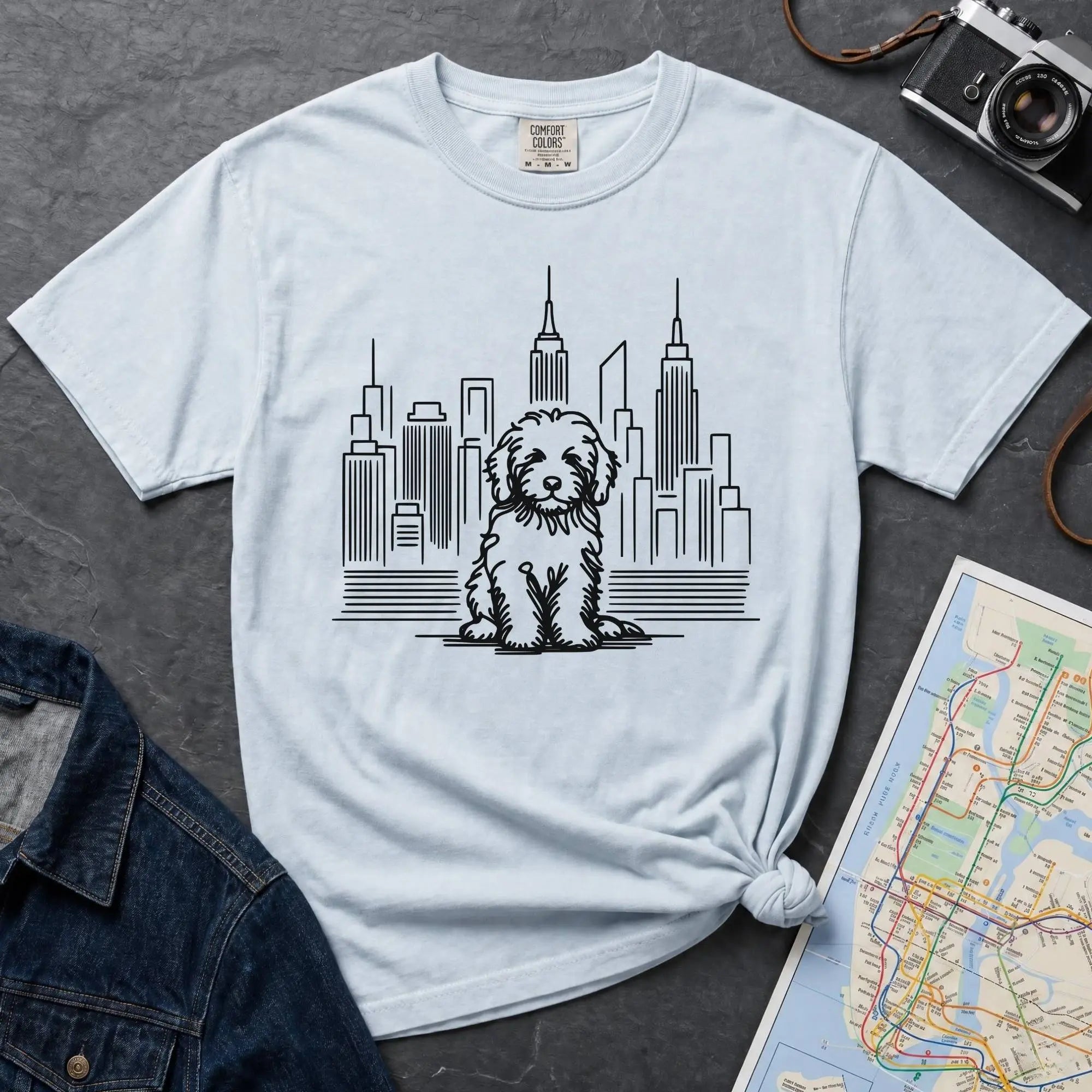 Light blue t-shirt with a Golden Doodle dog and cityscape design on a gray surface with a map and camera.