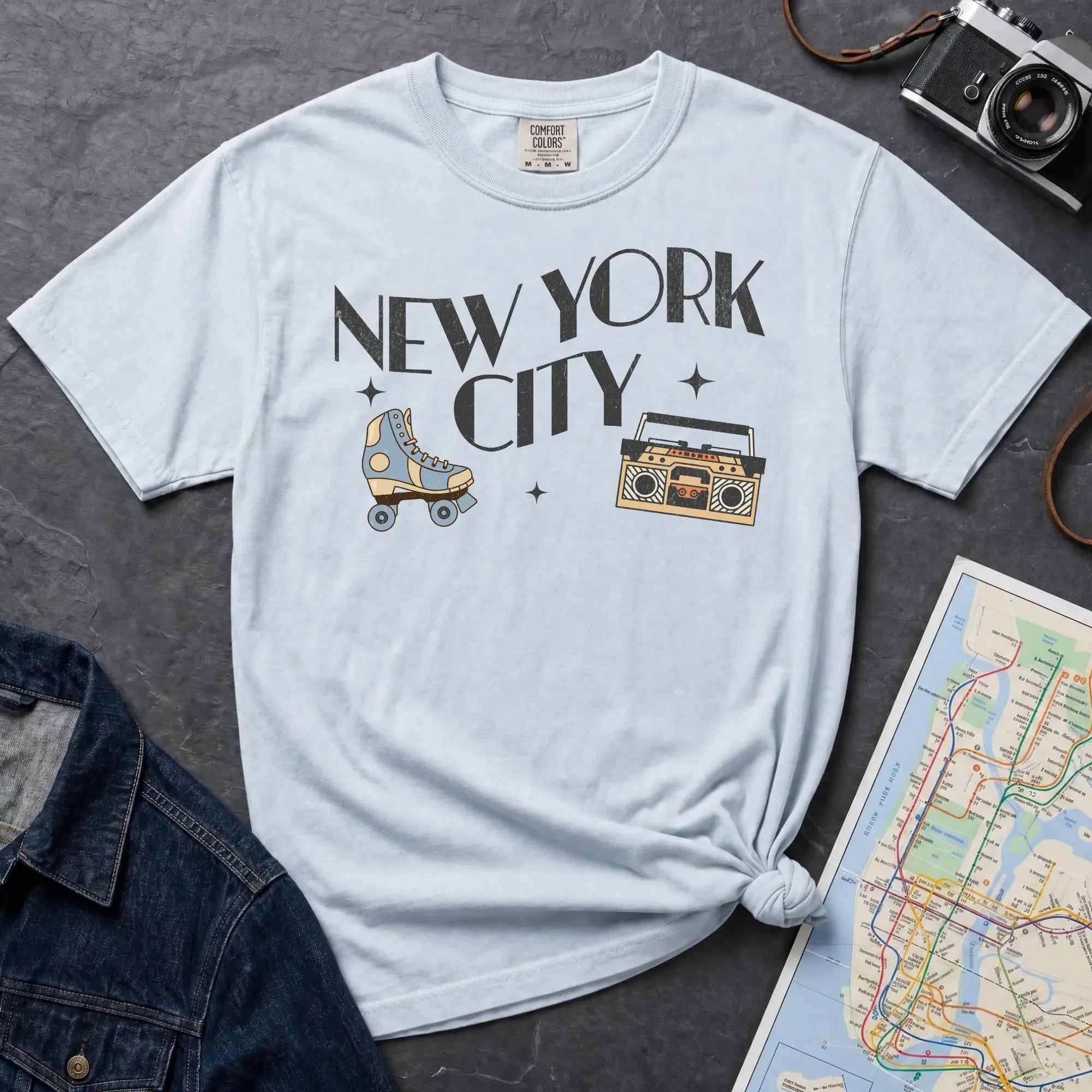 Light blue t-shirt with 'New York City' text and a roller skate and boom box on a dark surface with a map and camera.