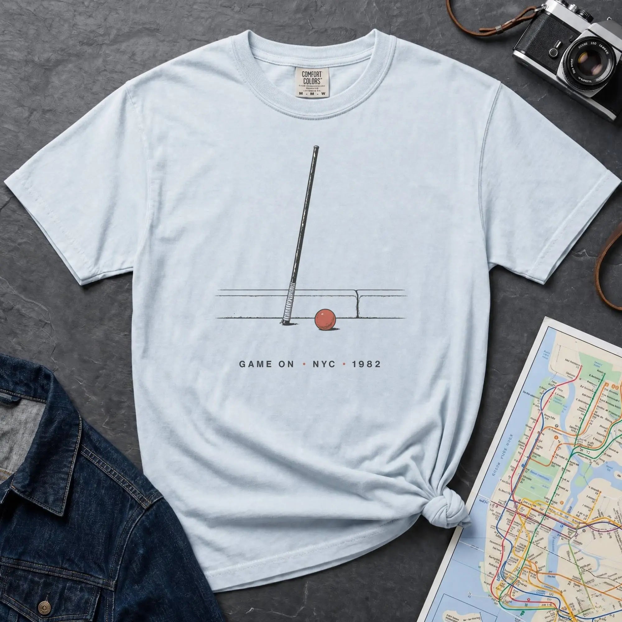 Light blue Comfort Colors t-shirt featuring a minimalist stickball bat and ball graphic with “Game On • NYC • 1982” text.