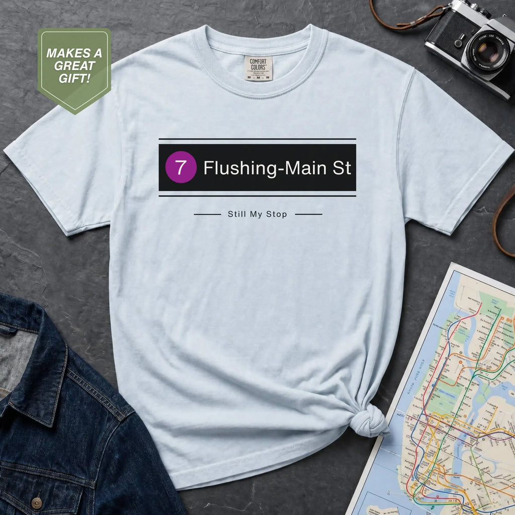 Light blue t-shirt with 'Flushing-Main St' design on a gray surface with a map and camera.