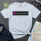 Light blue t-shirt with 'Flushing-Main St' design on a gray surface with a map and camera.
