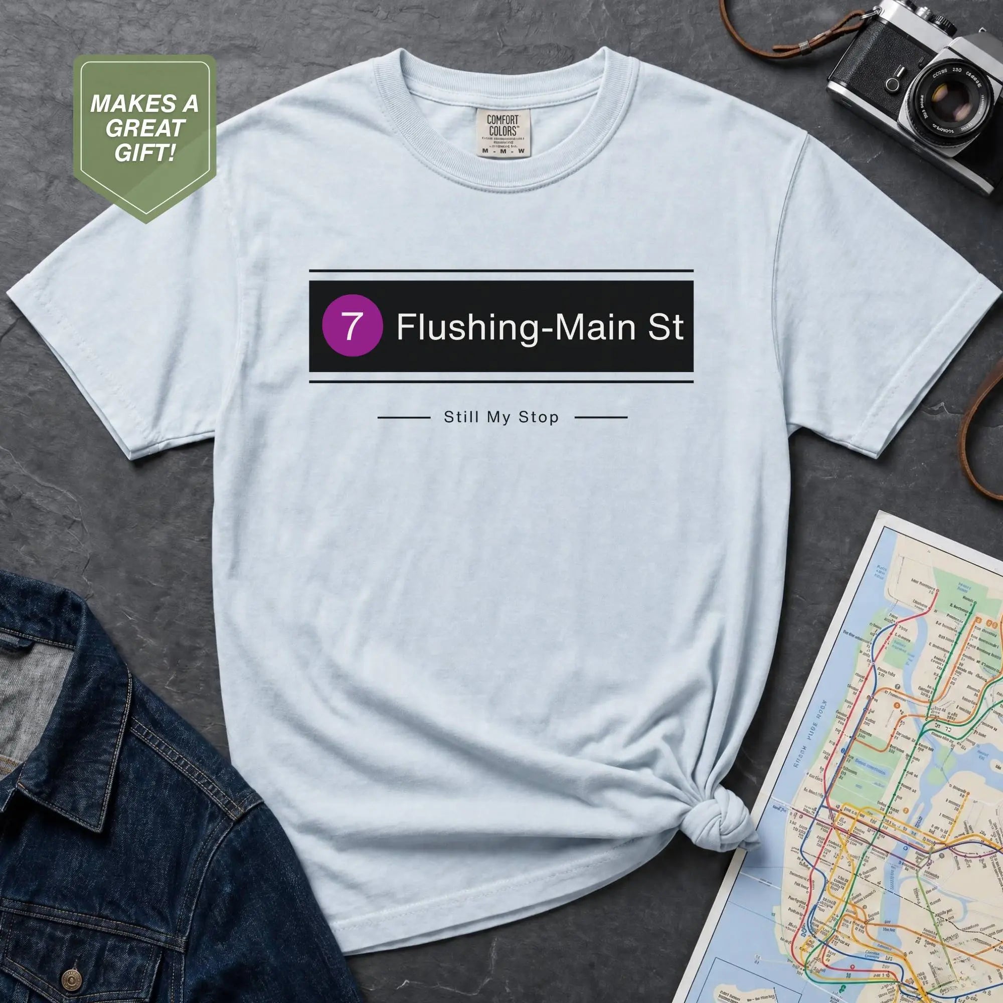 Light blue t-shirt with 'Flushing-Main St' design on a gray surface with a map and camera.