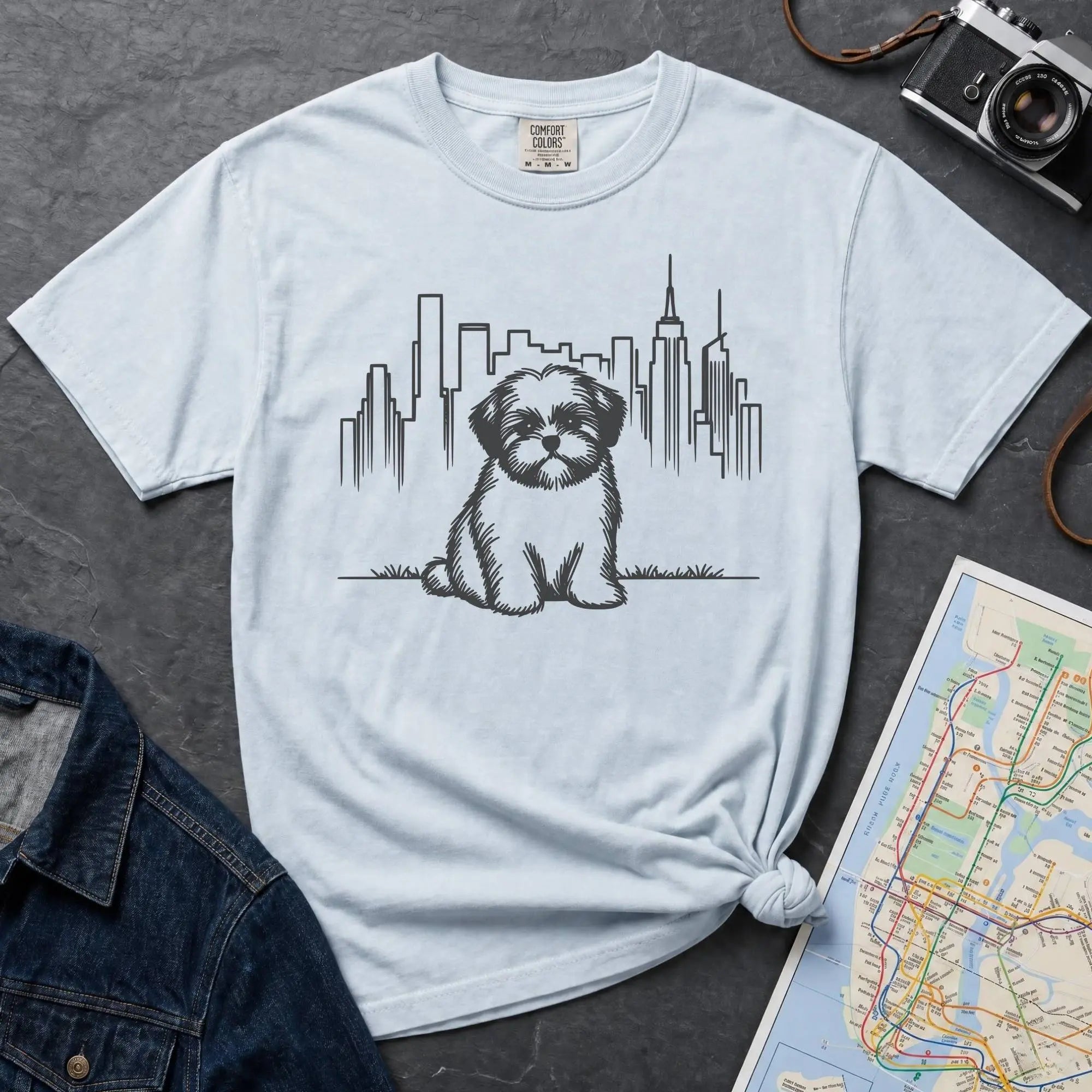 Light blue t-shirt with a Shih Tzu dog and cityscape design on a dark surface with a map and camera.