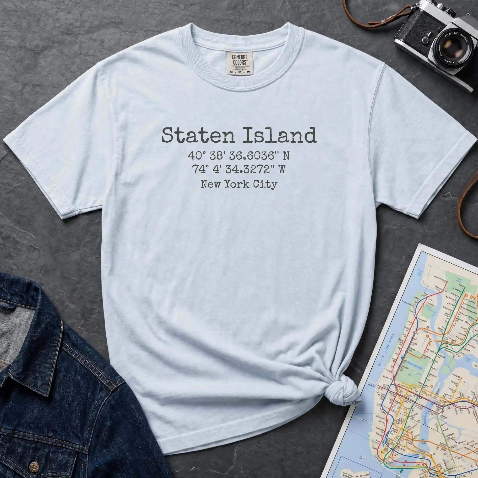 Light blue t-shirt with Staten Island text and coordinates on a dark surface with denim jacket and map.