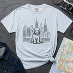 Light blue t-shirt with a Golden Doodle dog and cityscape design on a gray surface with a map and camera.