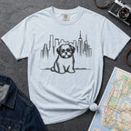 Light Blue t-shirt with a Shih Tzu dog and cityscape design on a dark surface with a map and camera.