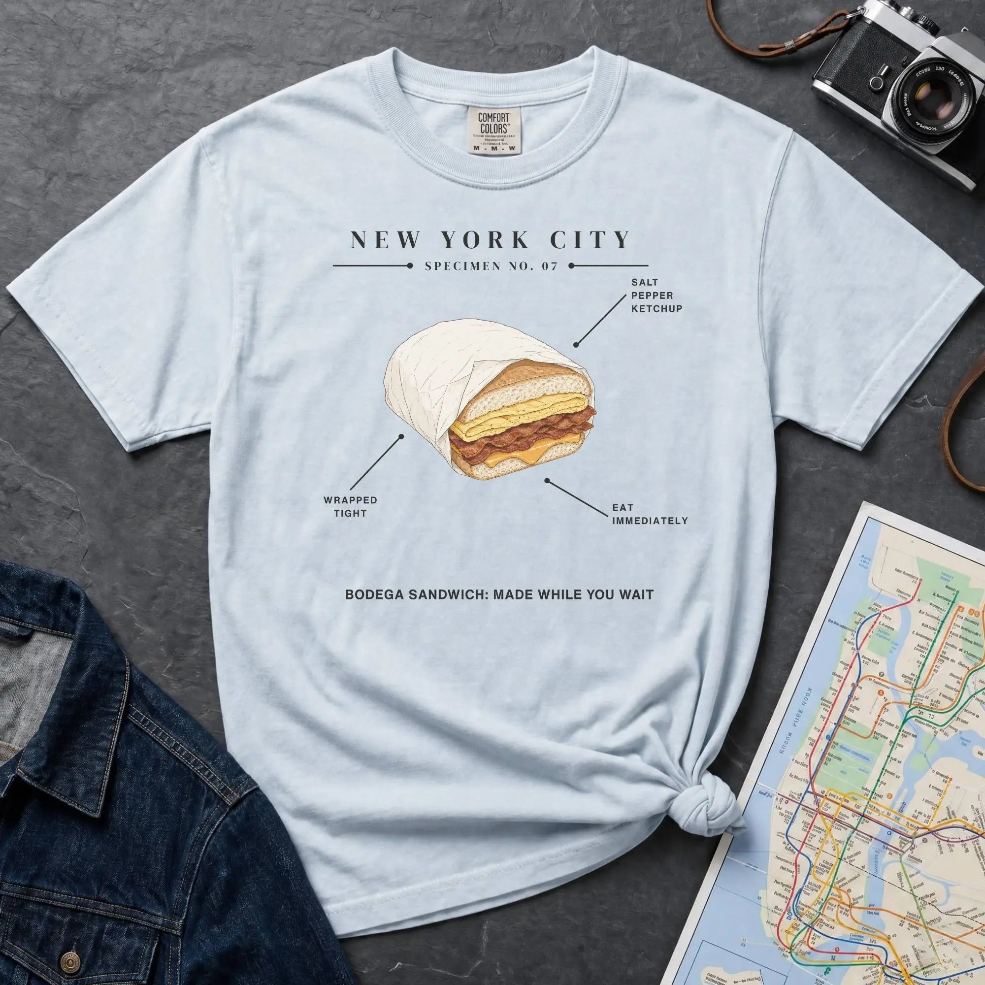 Light blue Comfort Colors t-shirt featuring a New York City bodega sandwich graphic with specimen-style callouts and “Bodega Sandwich: Made While You Wait” text.