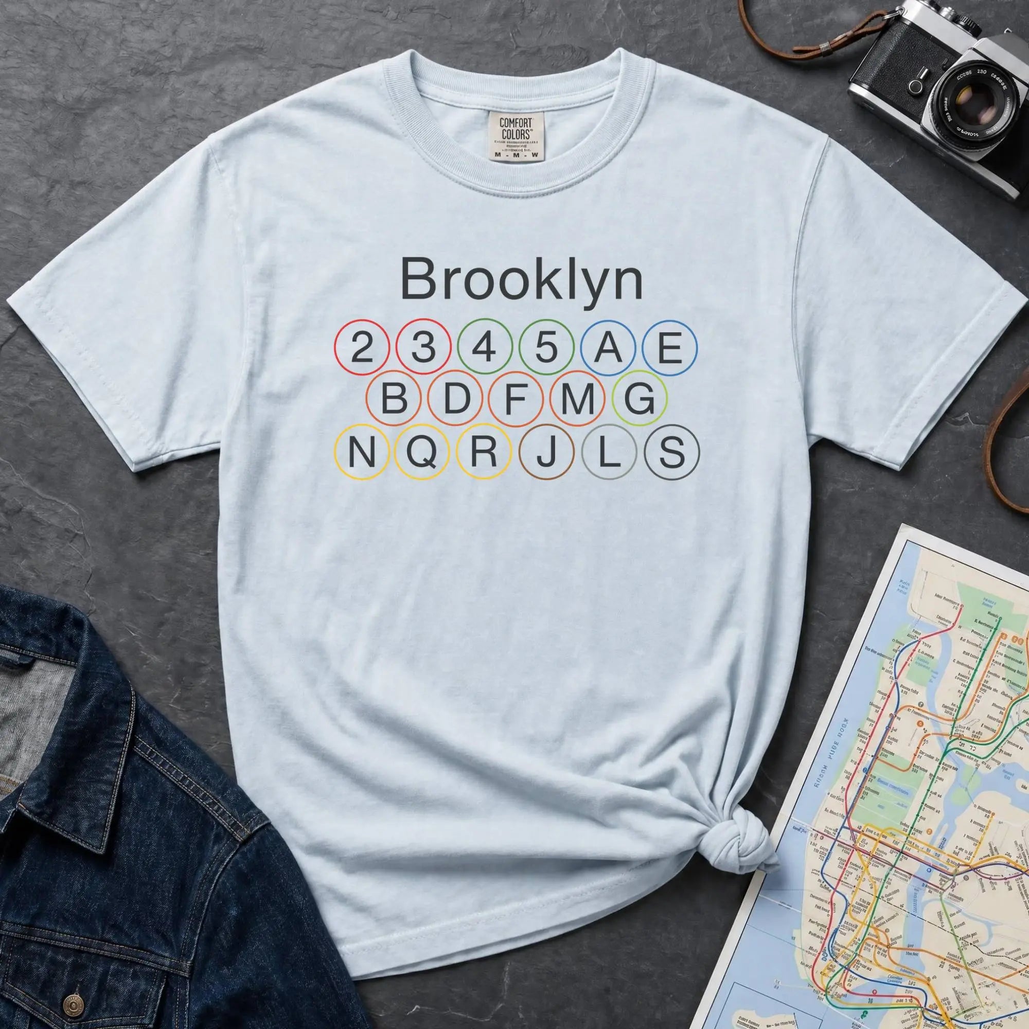 Light blue t-shirt with 'Brooklyn' and subway line numbers printed on it, placed on a dark surface with a map and denim jacket.