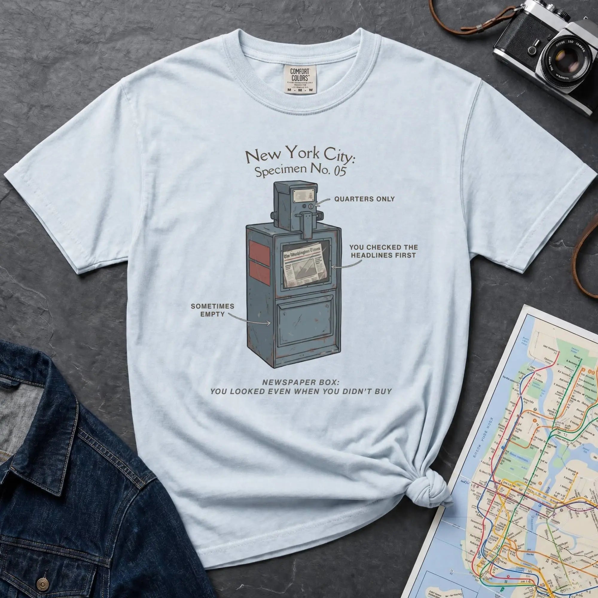 Light blue t-shirt with a graphic design of a newspaper box and text on a gray surface with a map and camera.