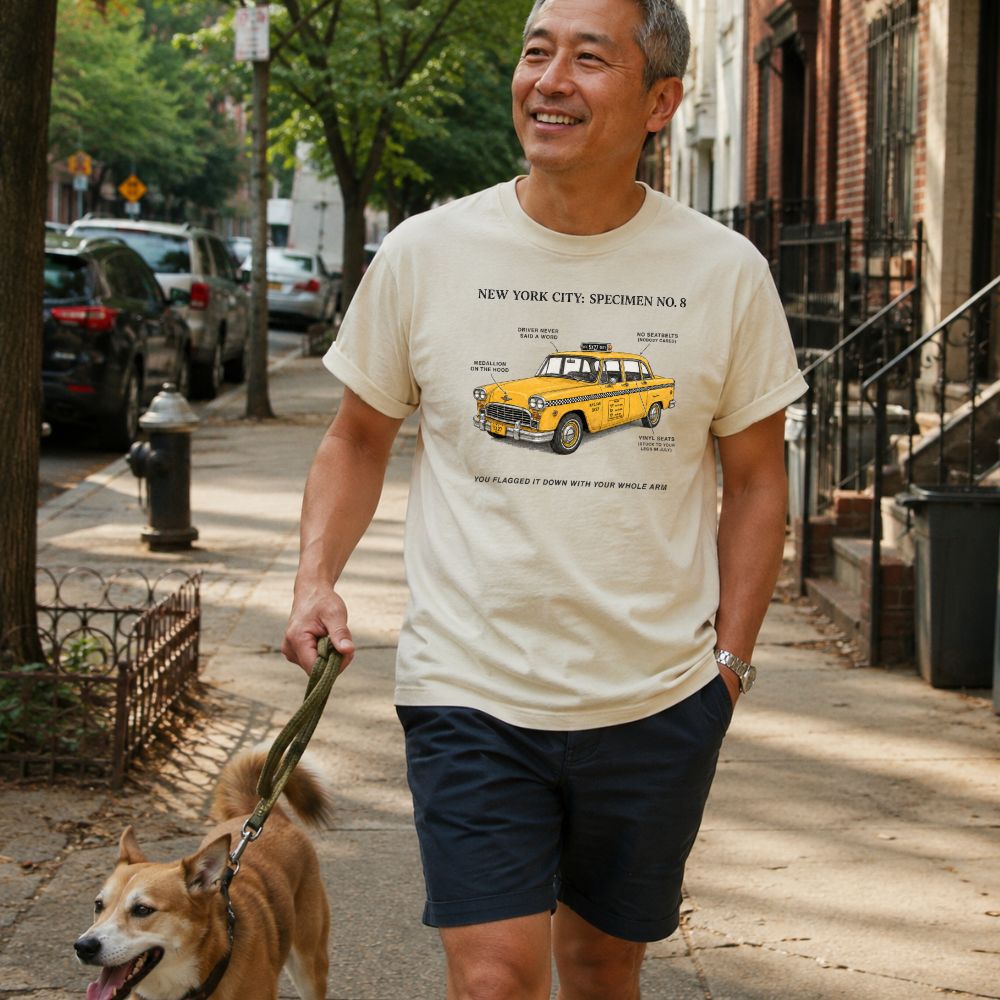 Man walking a dog on a sidewalk wearing an Ivory Comfort Colors t-shirt with yellow taxi graphic and text.